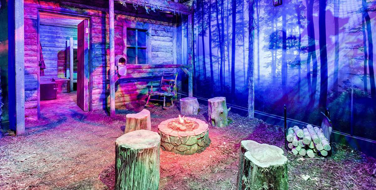 A cozy, illuminated cabin interior with a fire pit surrounded by logs, decorated with a forest mural on the walls.