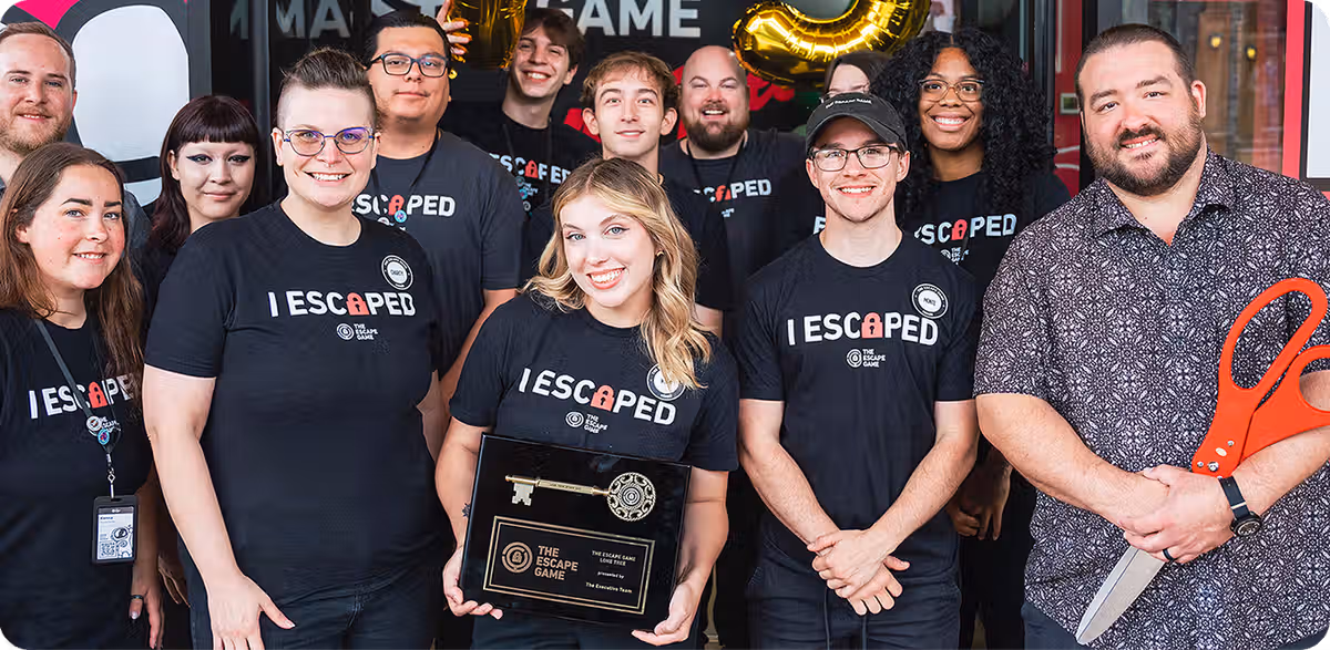 A group of employees wearing black shirts with "I ESCAPED" text stands together, smiling. One holds a trophy and another has scissors.
