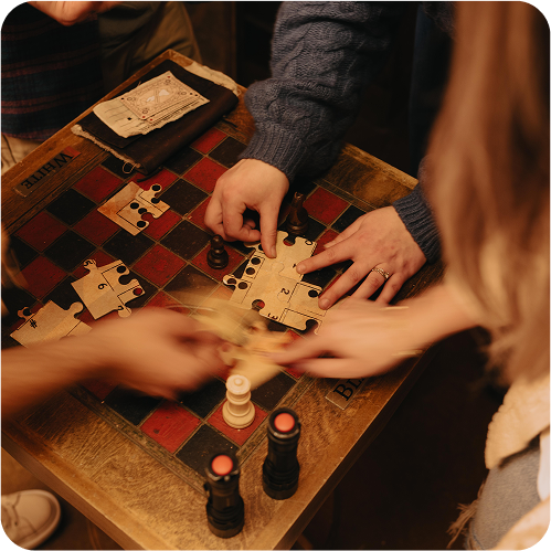 Hands reaching for puzzle pieces on a game board with chess pieces, flashlight stands, and a strategy note on the side.