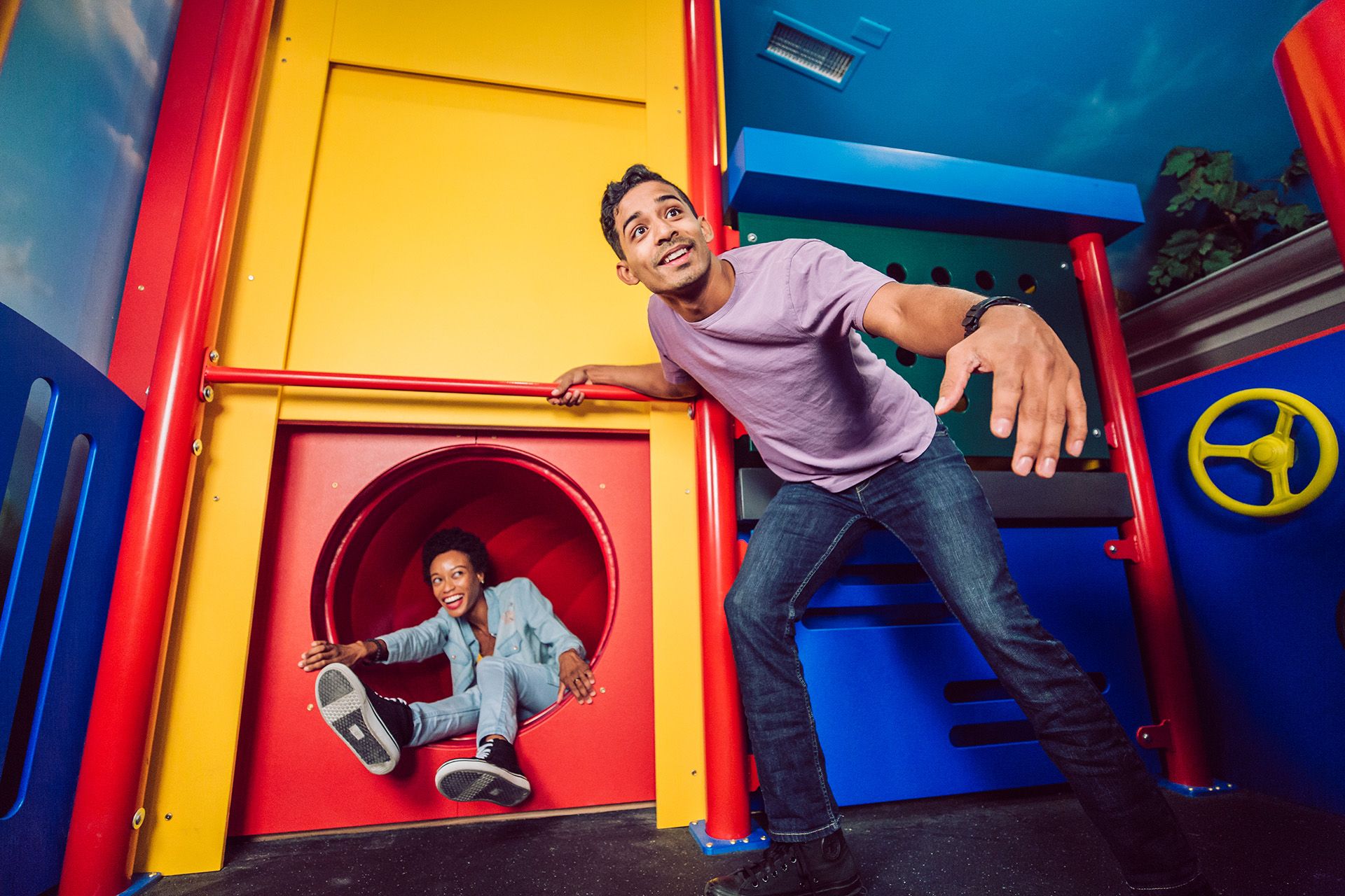 Two people play on a colorful playground structure. A person slides through a large tunnel while another reaches out.