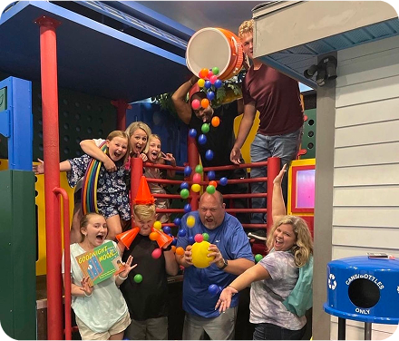 A group of eight people playfully expressing joy in a colorful indoor play area, with a bucket spilling balls.