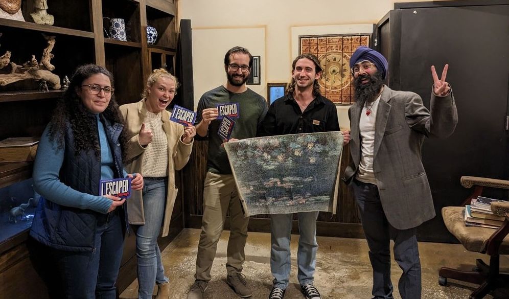 A group of five people stands in a cozy room, holding "I ESCAPED" signs. One person displays a piece of art.