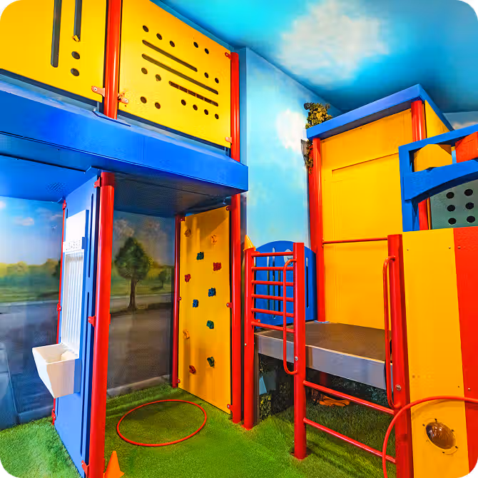 Colorful indoor play structure featuring climbing walls, slides, and interactive panels, set against a sky-themed background.