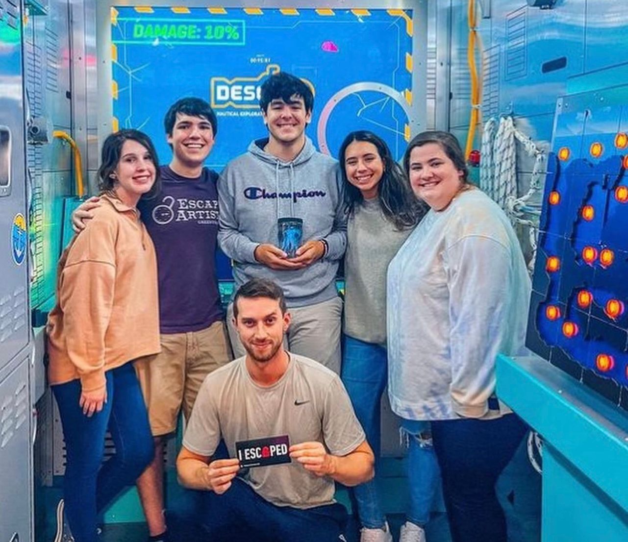 Group of six friends smiling in a themed escape room, holding a "I ESCAPED" sign, with colorful background elements.