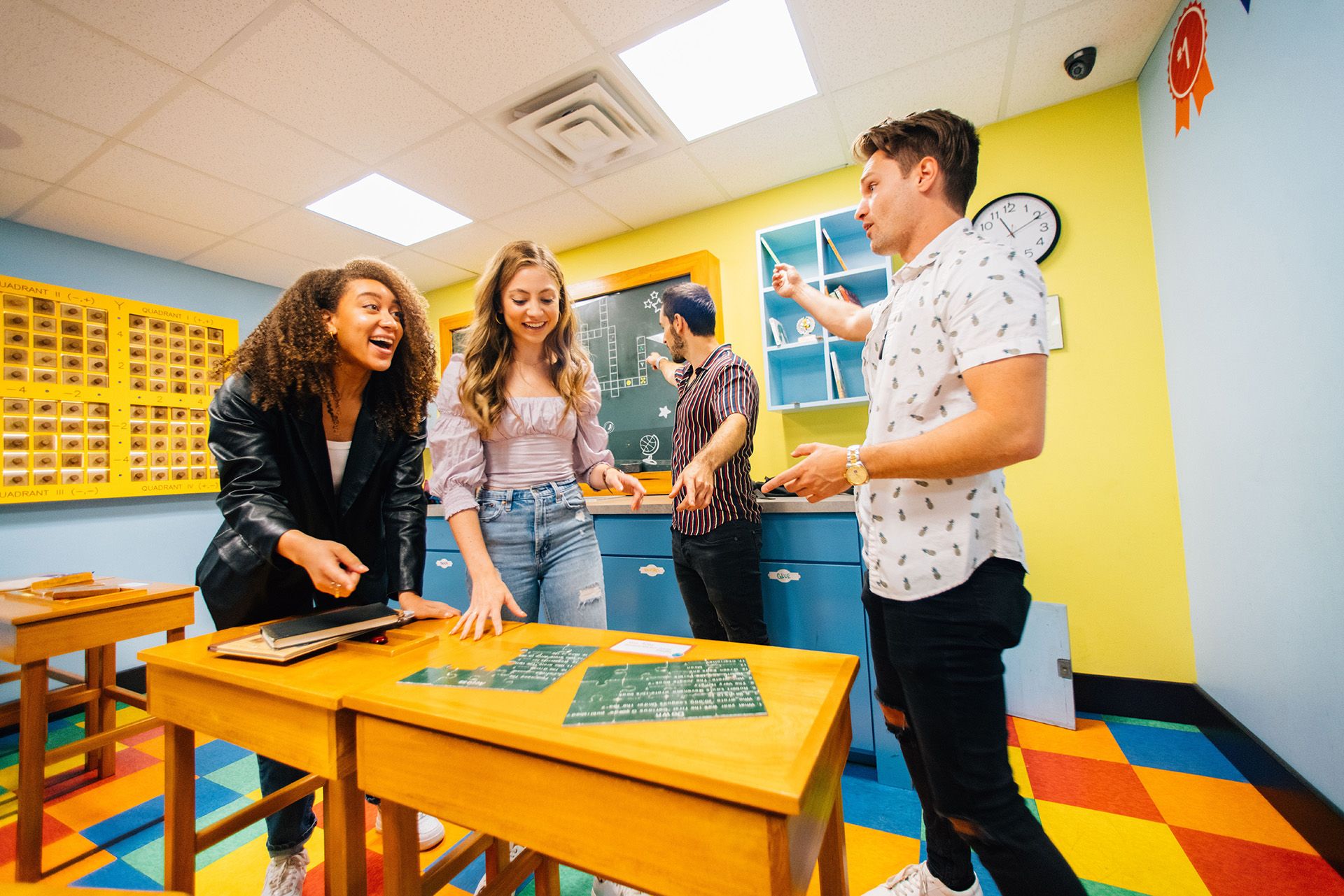 Four young adults are interacting in a colorful classroom, smiling and engaged. Two are at a table with notebooks, while the others stand nearby, discussing something on a chalkboard.