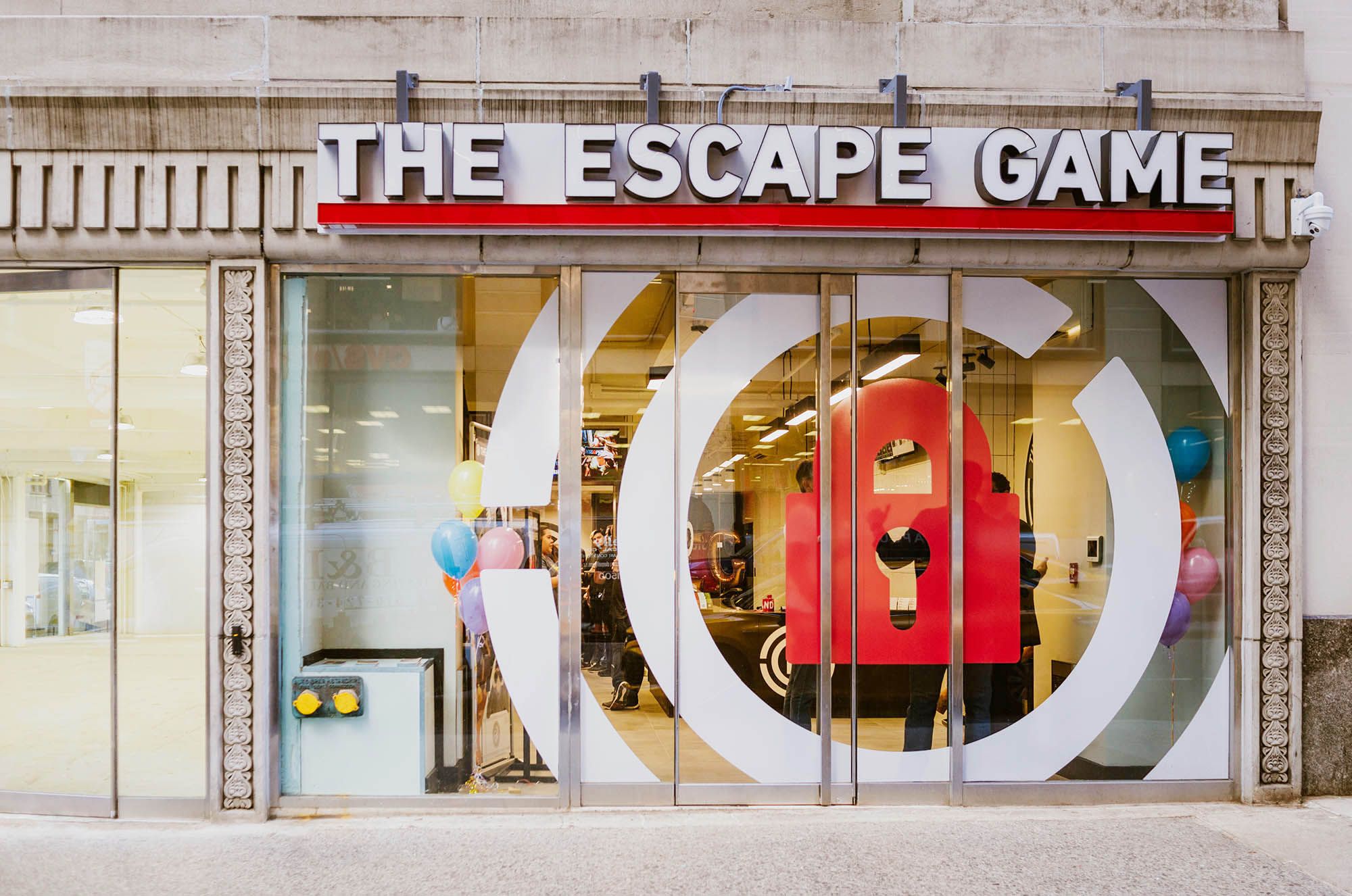 Storefront of "The Escape Game" with a large red lock graphic on glass doors and colorful balloons outside.