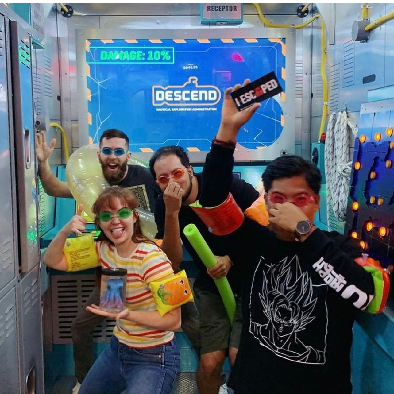 Four people posed playfully in an adventure room, wearing colorful goggles and floaties, holding props and laughing. A screen displays "DESCEND" and "DAMAGE: 10%."