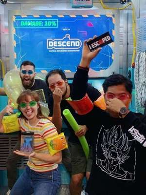 Four people posed playfully in an adventure room, wearing colorful goggles and floaties, holding props and laughing. A screen displays "DESCEND" and "DAMAGE: 10%."