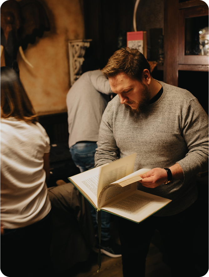 A man in a gray sweater browses through a book, with two people visible in the background. Vintage decor is present.