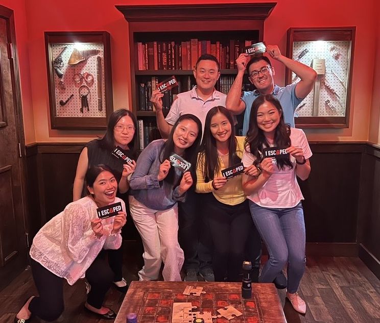 Group of eight smiling people inside a dimly lit room, holding "I ESCAPED" signs. A wooden table and shelves filled with books and tools are in the background.