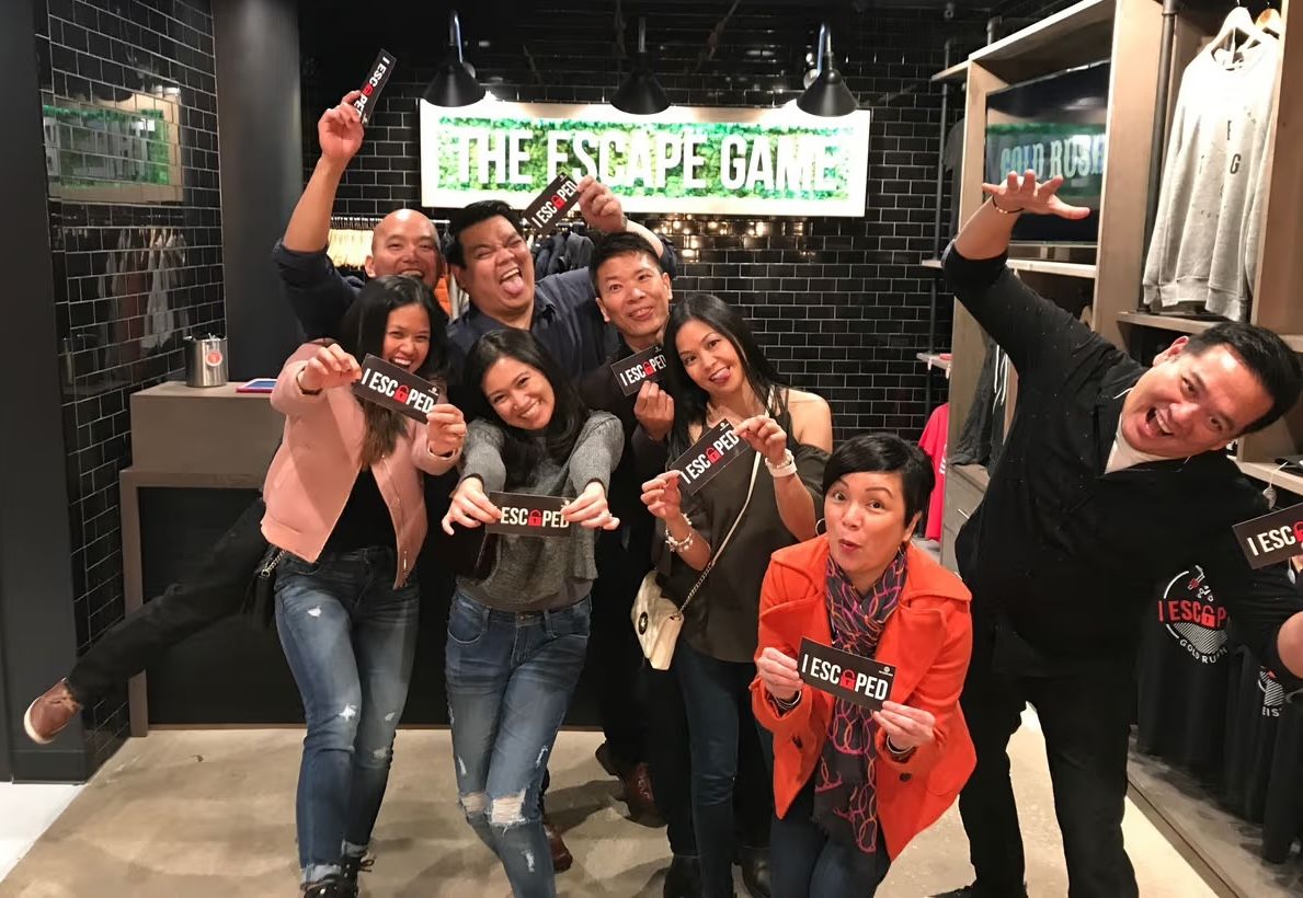 Group of eight people posing with playful expressions, holding signs that say "I ESCAPED" in a store setting.