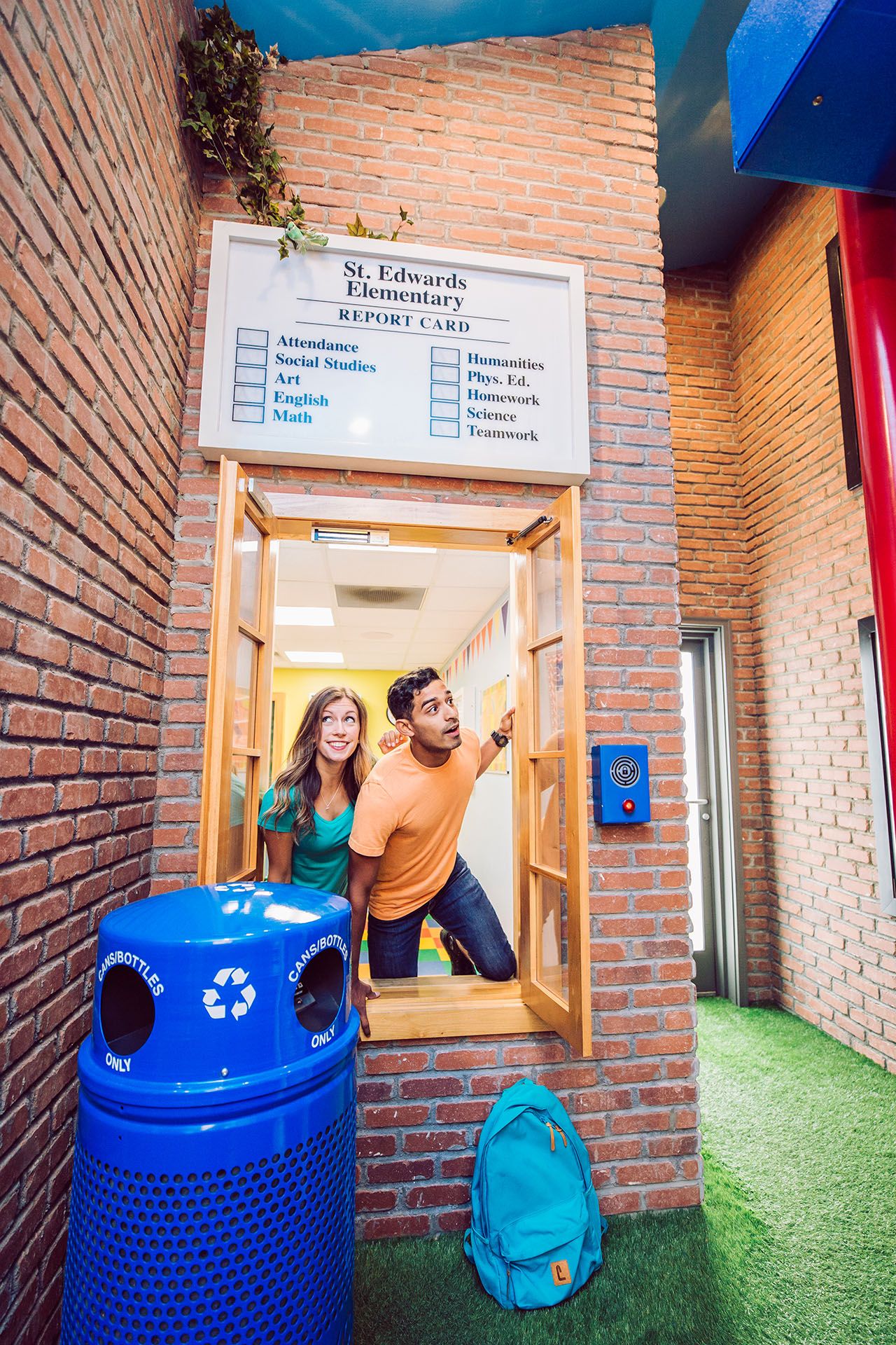 Two students, a girl with long hair in a teal shirt and a boy in an orange shirt, peek through a window. Behind them is a brick wall with a report card sign listing subjects. A blue recycling bin is in the foreground, with a teal backpack beside it.