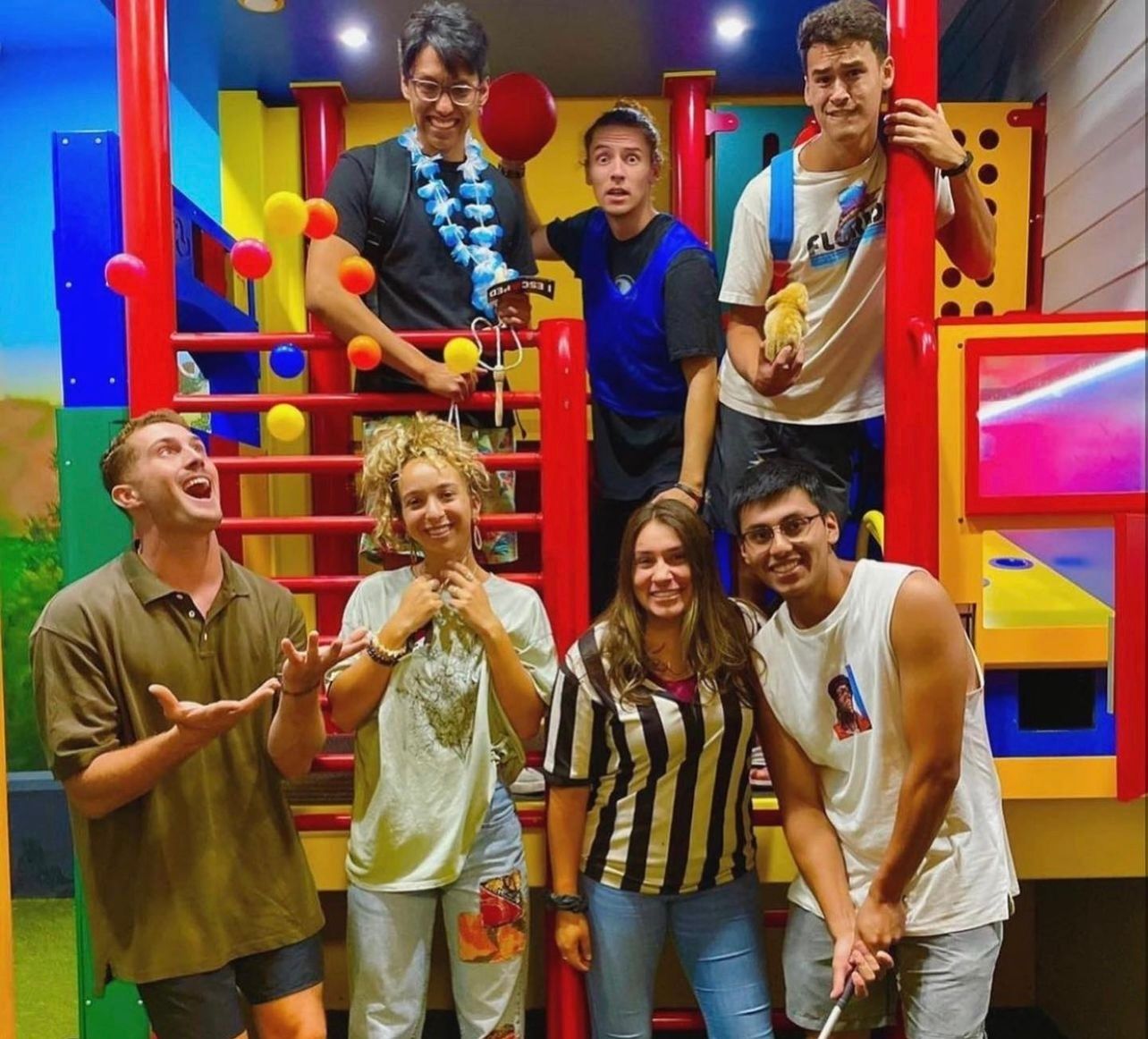 Group of seven friends posing in a colorful play area, smiling, with one person juggling balls and others holding props.