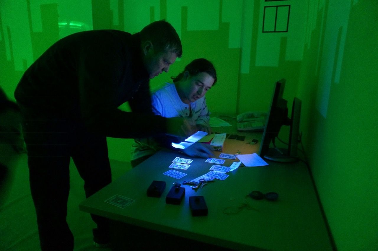 Two people in a green-lit room examine cards and documents on a table, with one using a phone to illuminate the items.