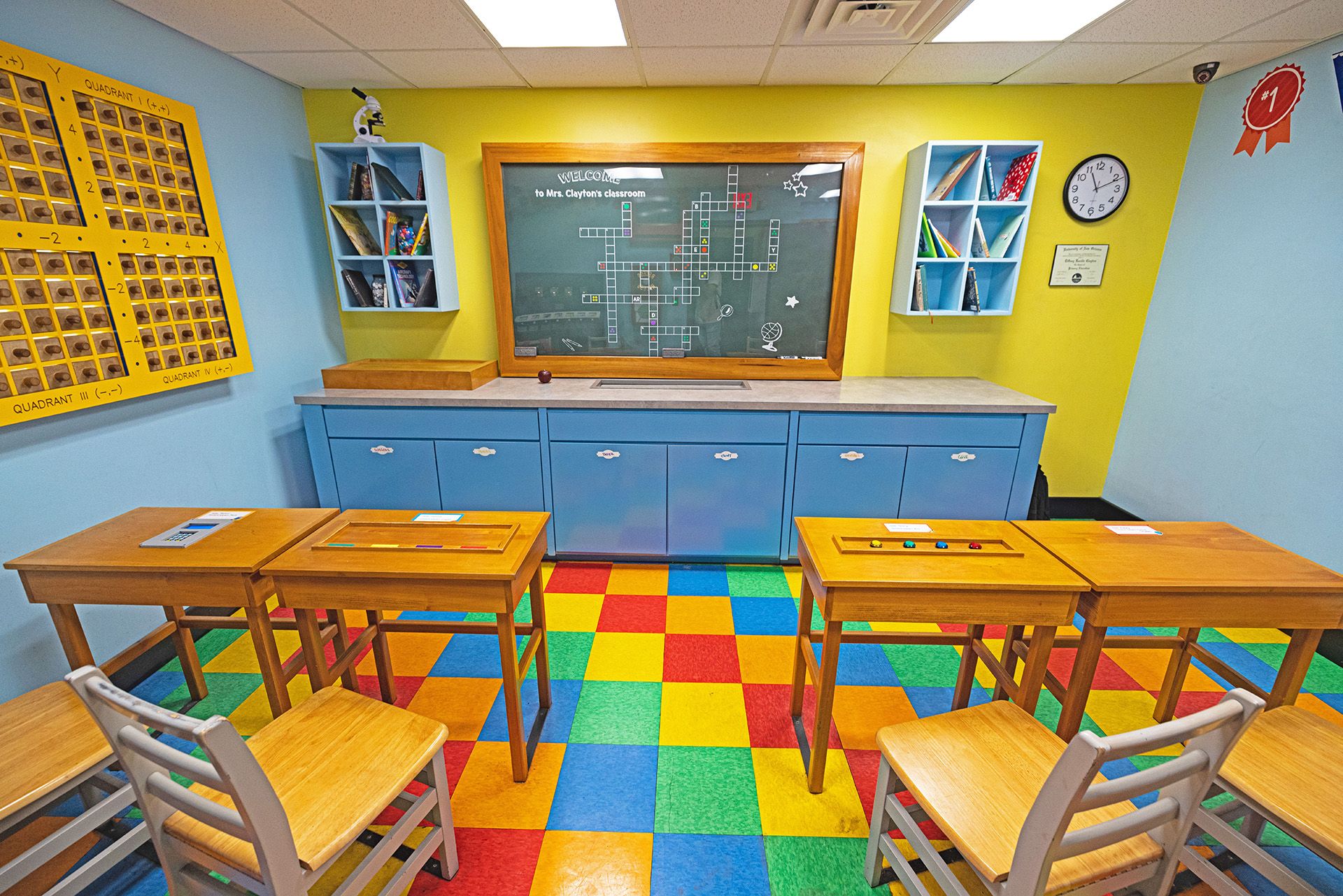 A colorful classroom with desks facing a chalkboard, shelves of books, and a math poster on the wall.
