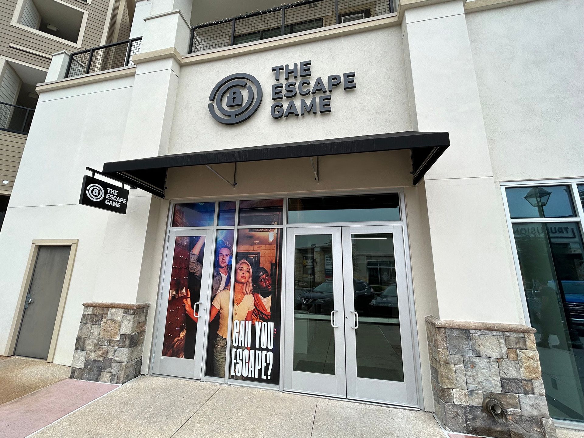 Entrance of "The Escape Game" featuring double glass doors with a sign above and a promotional image of players.