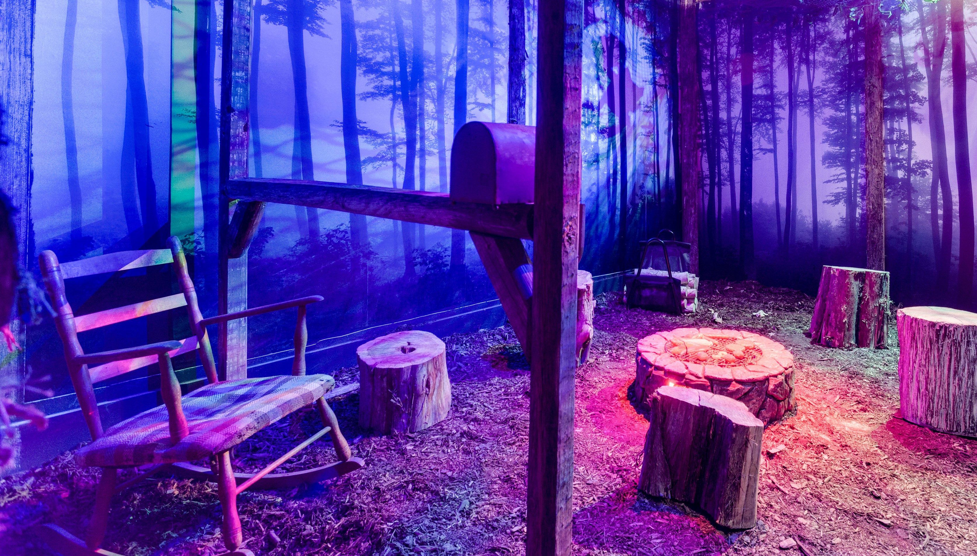 A rustic setting with a rocking chair, mailbox, and tree stumps on a forest-themed backdrop lit in purple and blue tones.