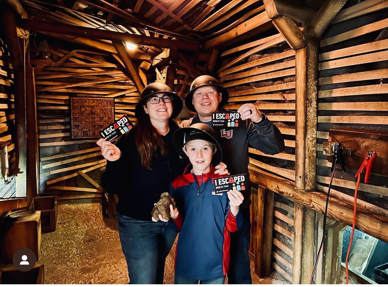 Three people stand inside a wooden room, holding up "I ESCAPED" cards. The woman wears glasses and a black hat, the man wears a gray shirt, and the child wears a red and blue jacket.