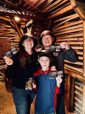 Three people stand inside a wooden room, holding up "I ESCAPED" cards. The woman wears glasses and a black hat, the man wears a gray shirt, and the child wears a red and blue jacket.