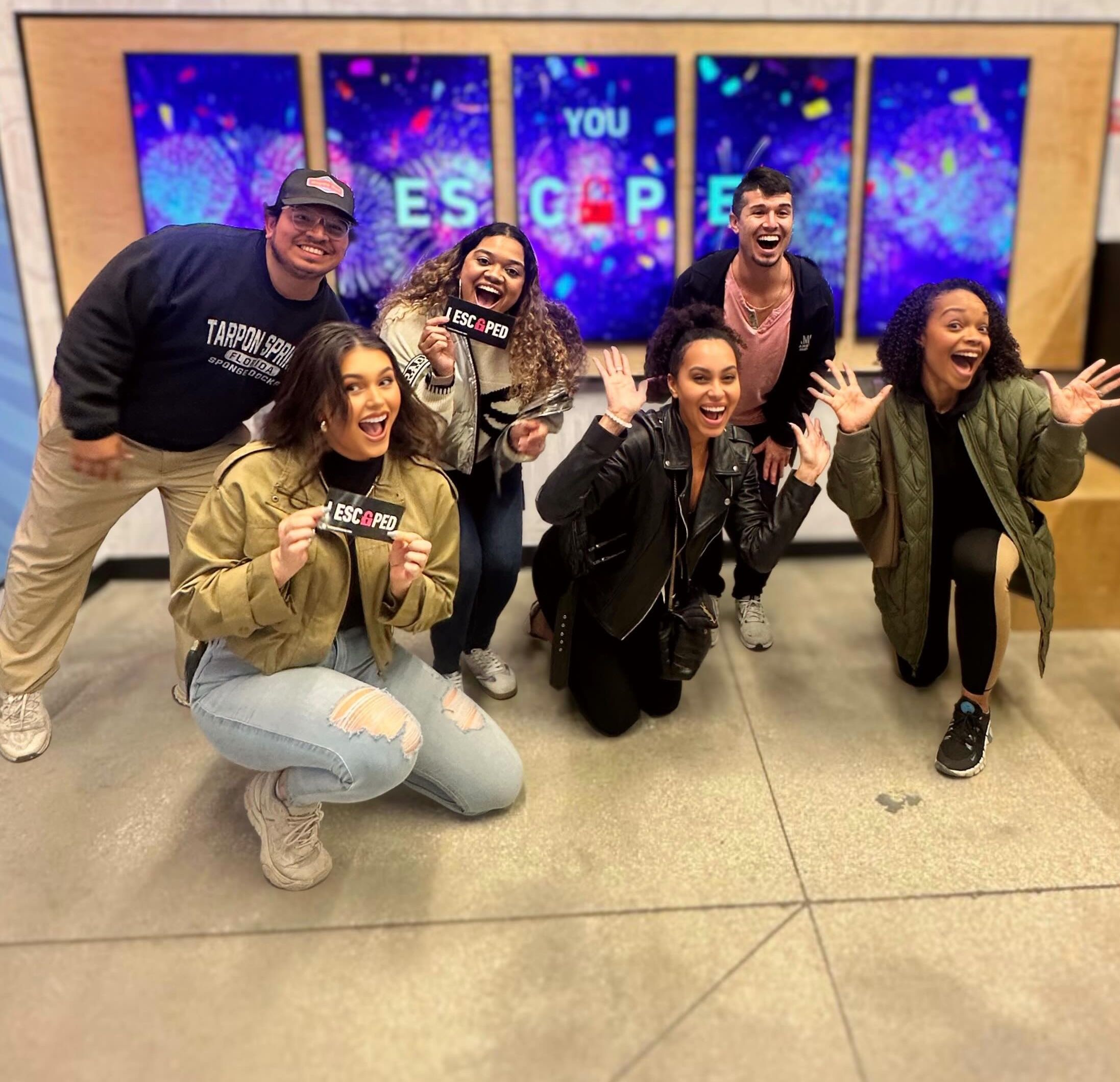 A group of five smiling friends poses together, holding an "ESCAPED" sign. Colorful screens with confetti are in the background.
