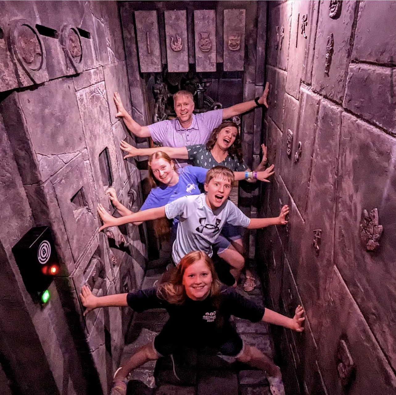 A group of five people poses in a narrow, dimly lit corridor with stone-like walls, all smiling and stretching out their arms.