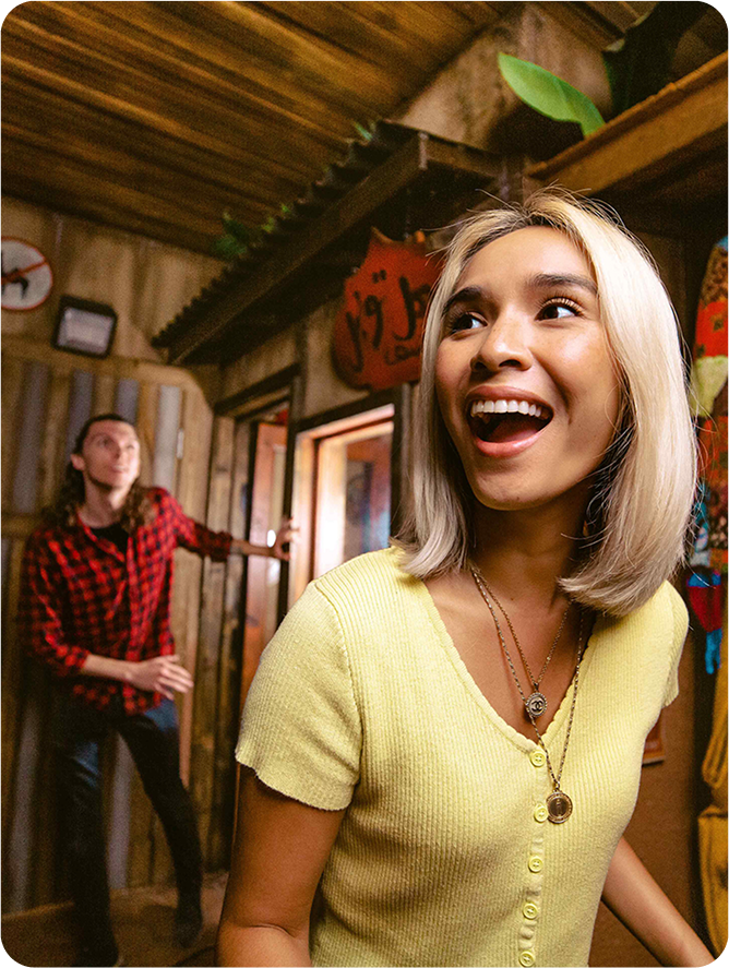 A woman with blonde hair smiles joyfully in a cozy interior, while a man in a red plaid shirt stands in the background.