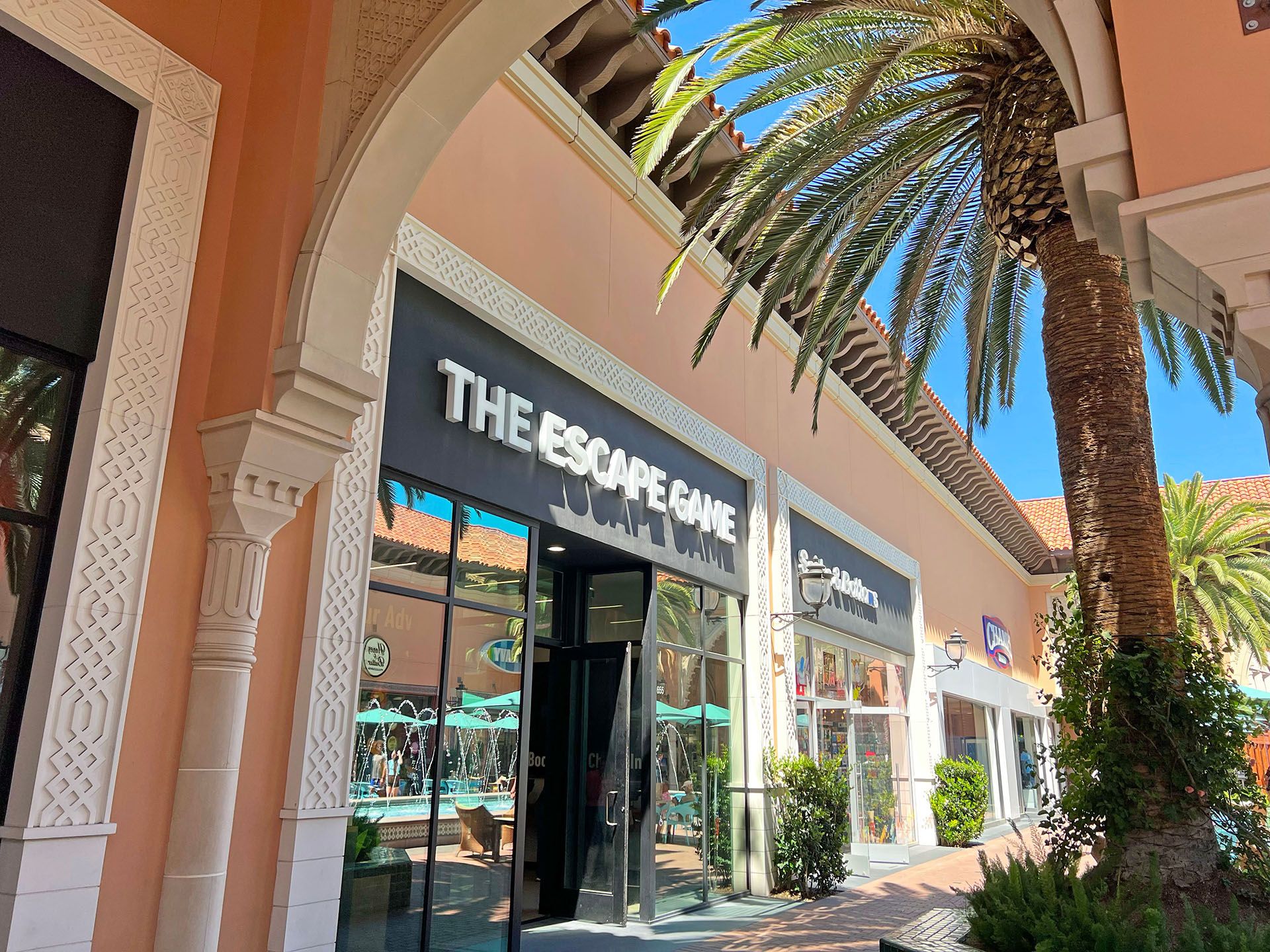 Storefront of "The Escape Game" with palm trees nearby and bright blue sky above; decorative architectural details visible.