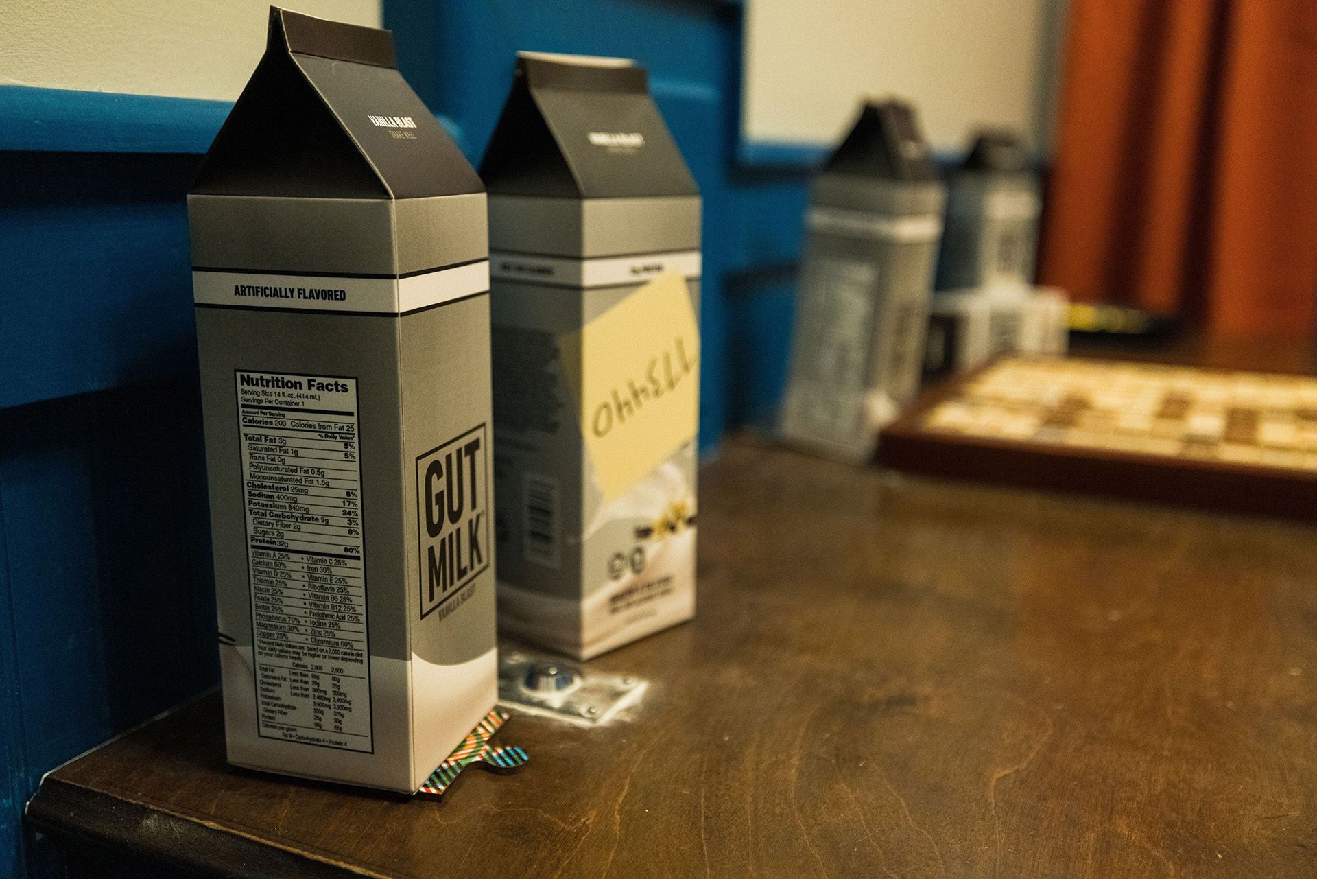 Three milk cartons labeled "GUT MILK" and "OhhSILL" sit on a wooden table, with a scrabble board partially visible in the background.