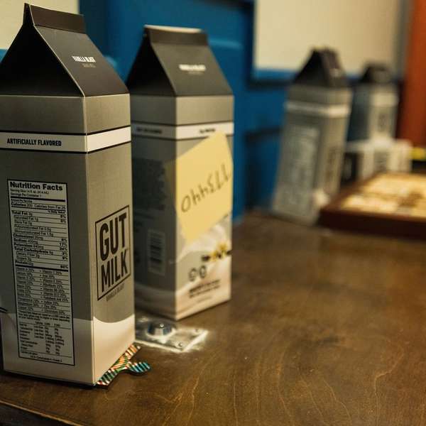 Three milk cartons labeled "GUT MILK" and "OhhSILL" sit on a wooden table, with a scrabble board partially visible in the background.