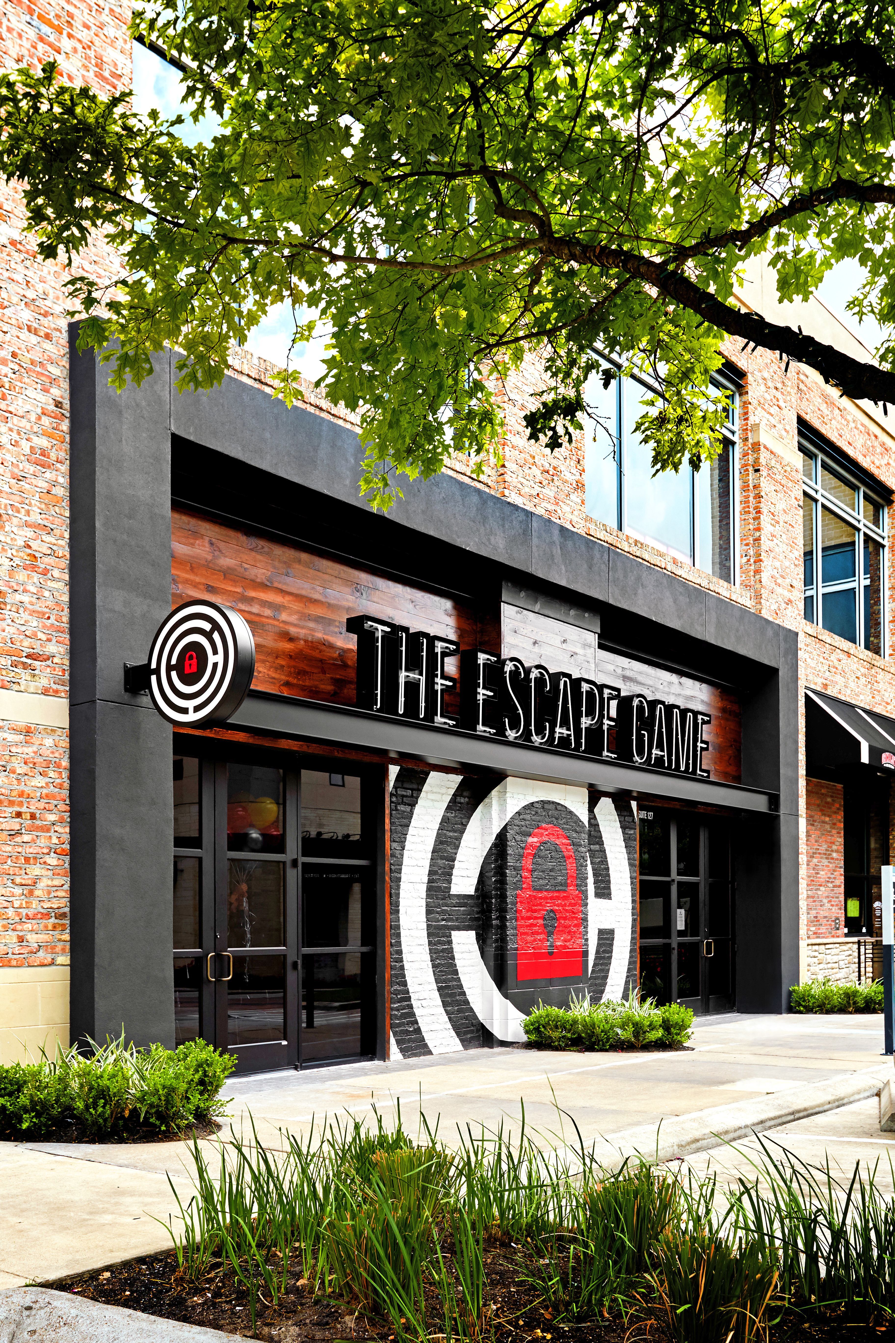 Exterior of "The Escape Game" building featuring a lock graphic on the wall, surrounded by greenery and urban architecture.