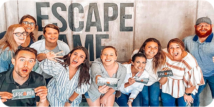 A group of ten people smiling and holding "I ESCAPED!" cards, standing in front of a large wall with the words "ESCAPE ME."
