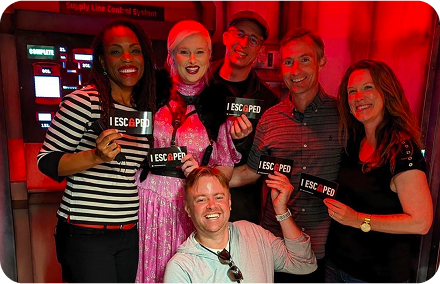 A group of six people, smiling and holding "I ESCAPED" signs, stand in a dimly lit room with a sci-fi backdrop.