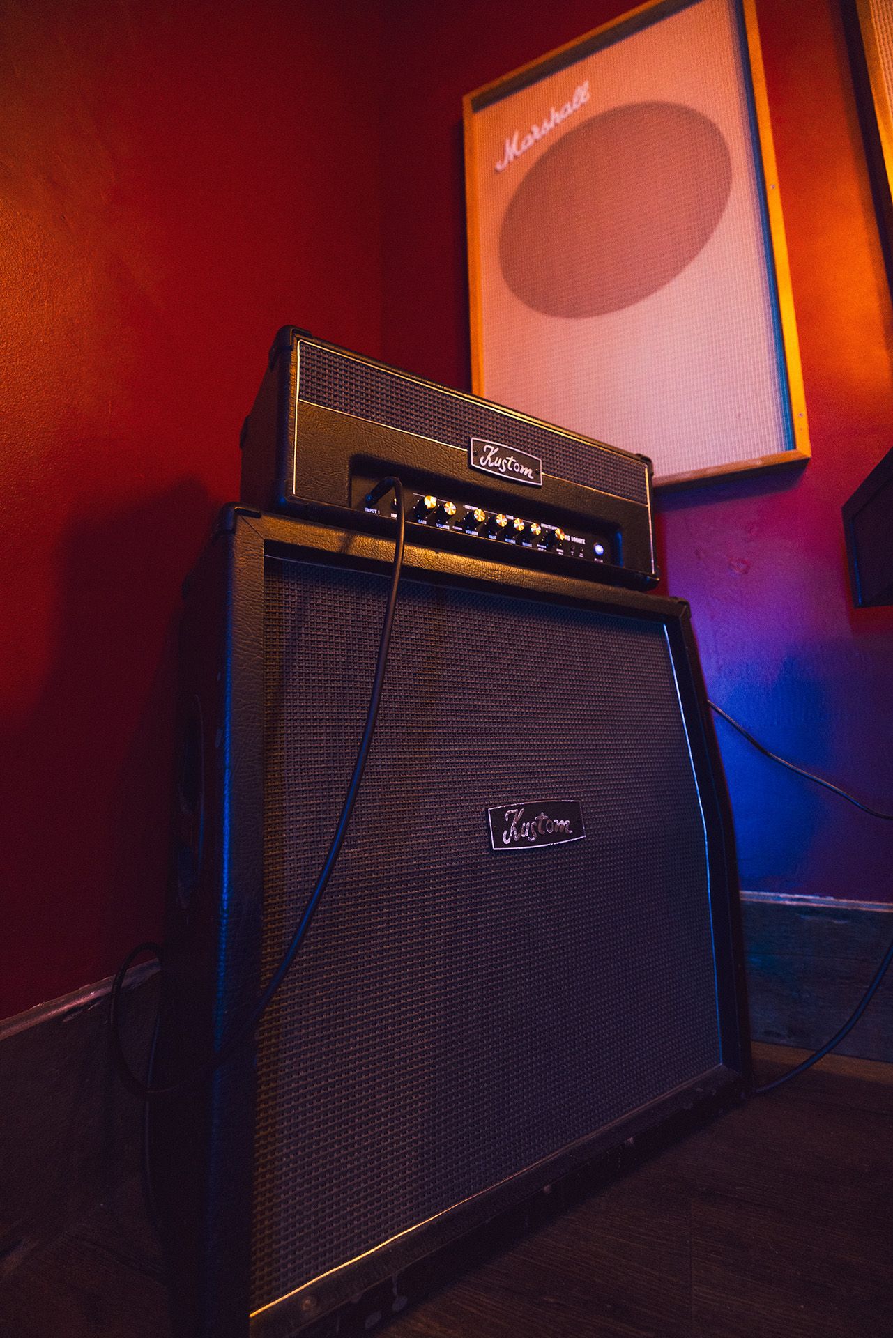 A Kustom guitar amplifier stacked with a speaker cabinet against a red wall, illuminated with blue lighting.