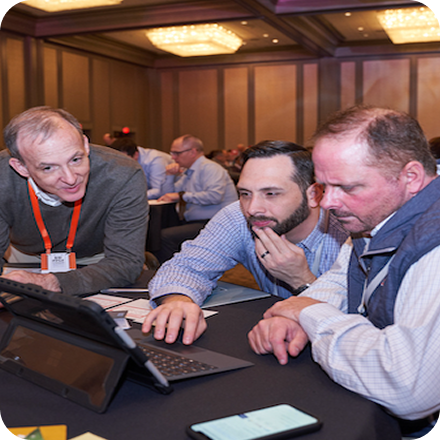 Three men are gathered around a laptop, focusing intently on the screen. The setting is a conference room.