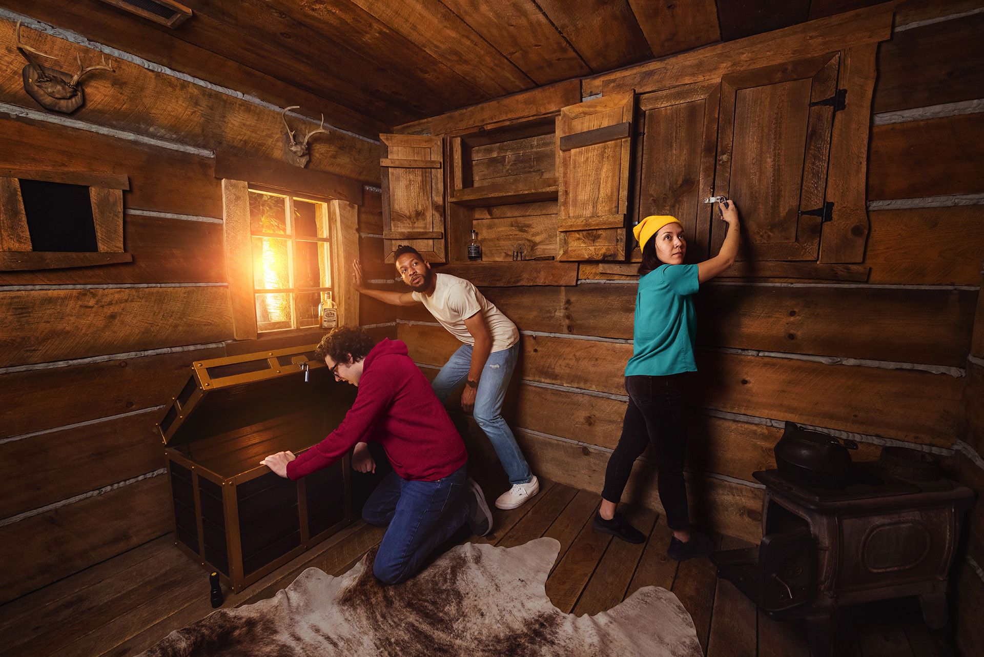 Three people are inside a rustic wooden cabin. One person is examining a treasure chest, while another is working on a cupboard, and the third is standing near a window with sunlight streaming in.