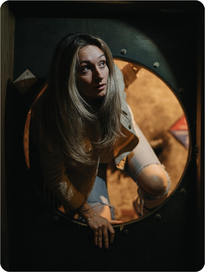 A woman with long hair is peeking out from a circular opening, looking upward with a concerned expression. She is wearing a light jacket and distressed jeans.