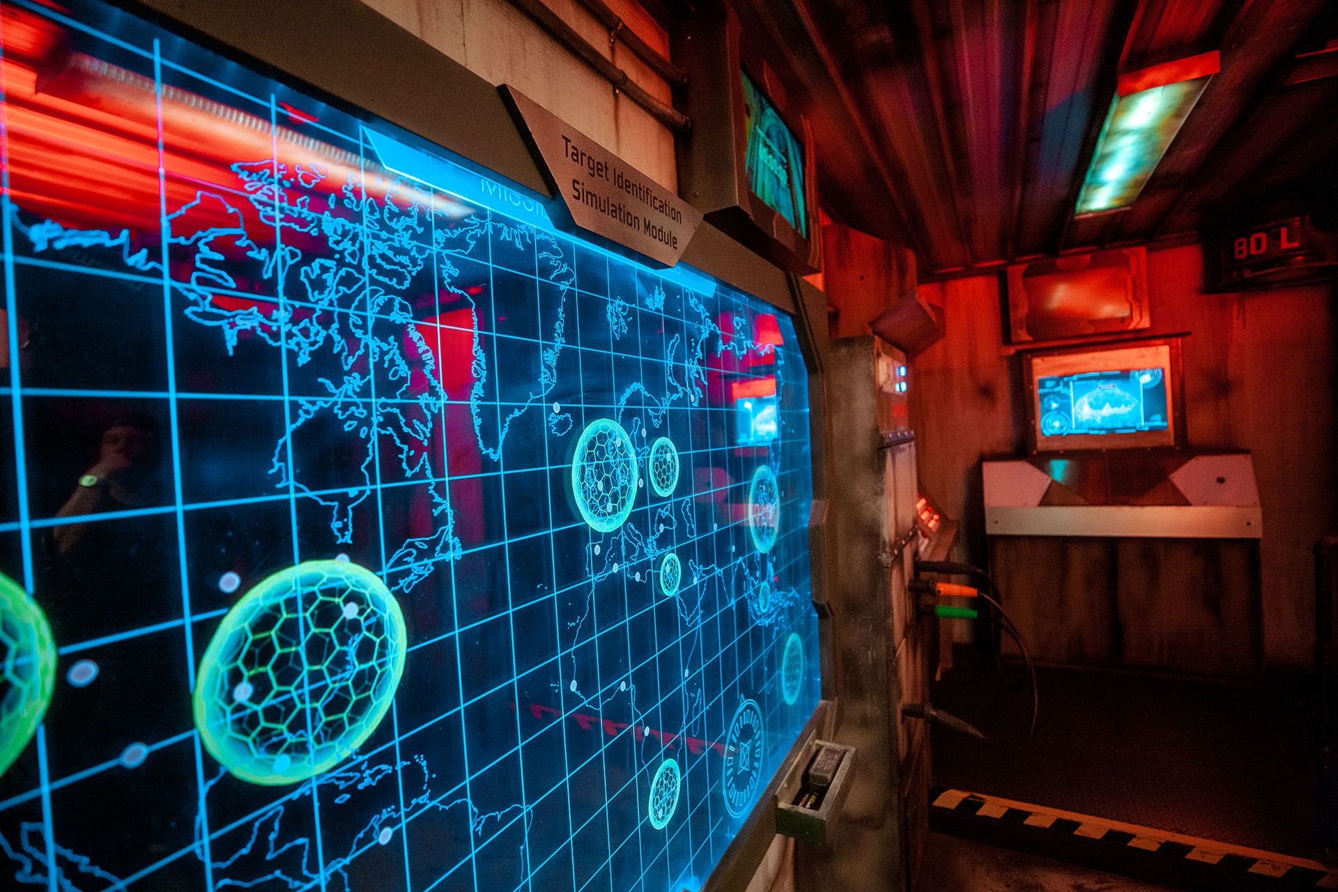 A glowing blue touchscreen map displays various locations and circular targets in a dimly lit room with colorful lights.