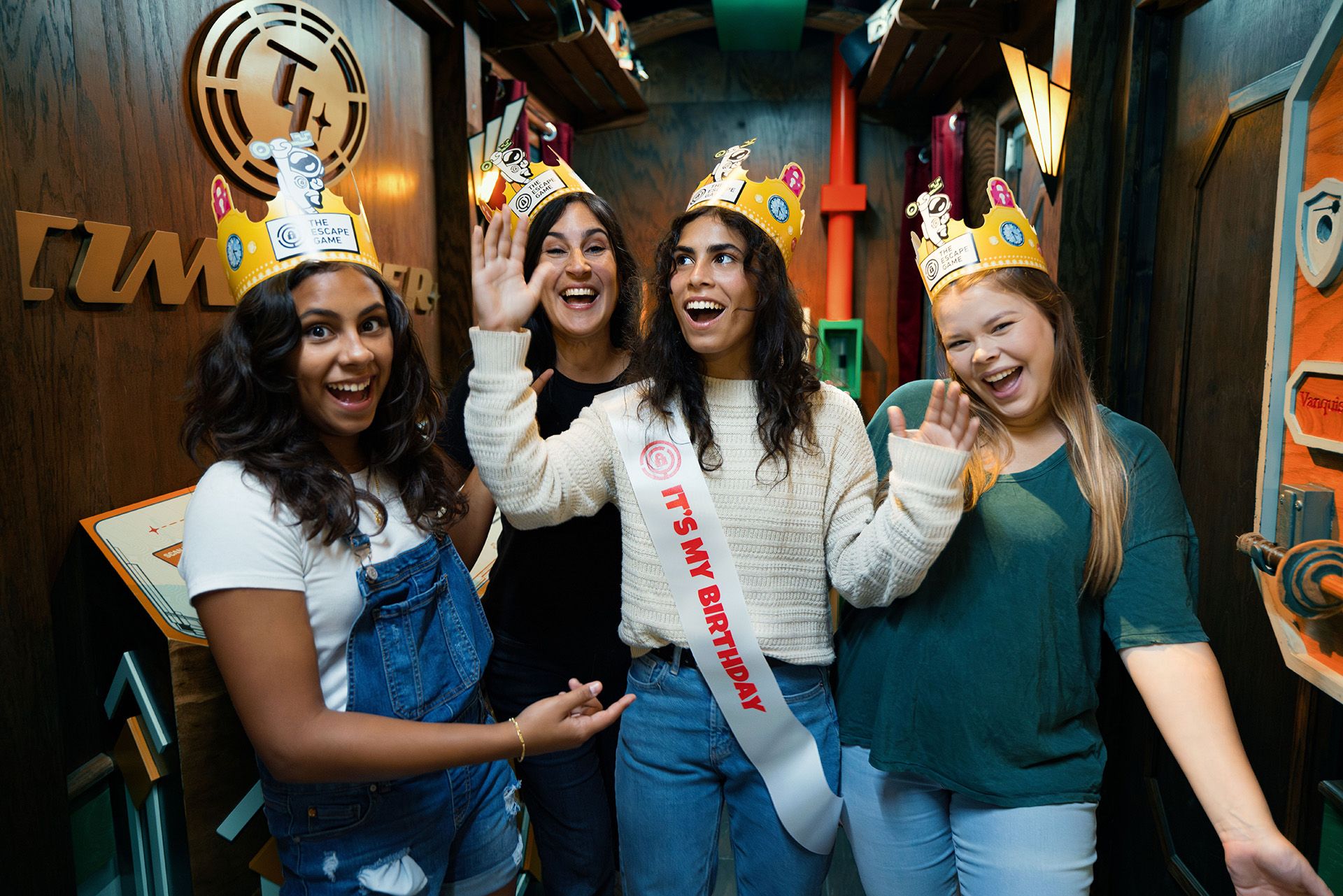 Four women celebrate a birthday in a colorful escape room, all wearing crowns. One woman holds a "It's My Birthday" sash.