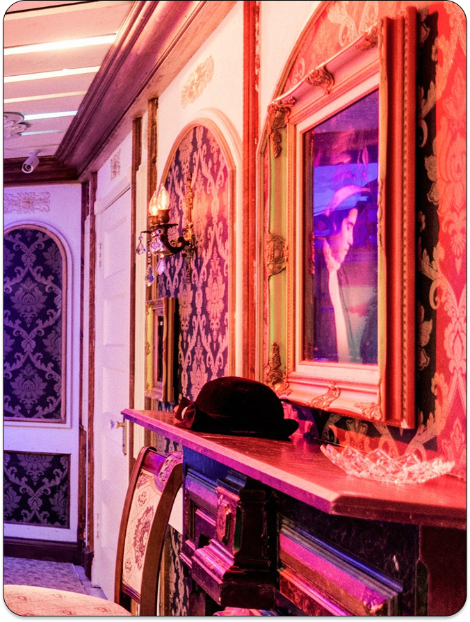 A decorated hallway with vintage wallpaper, a framed artwork, and a black hat resting on a mantel. Warm ambient lighting.
