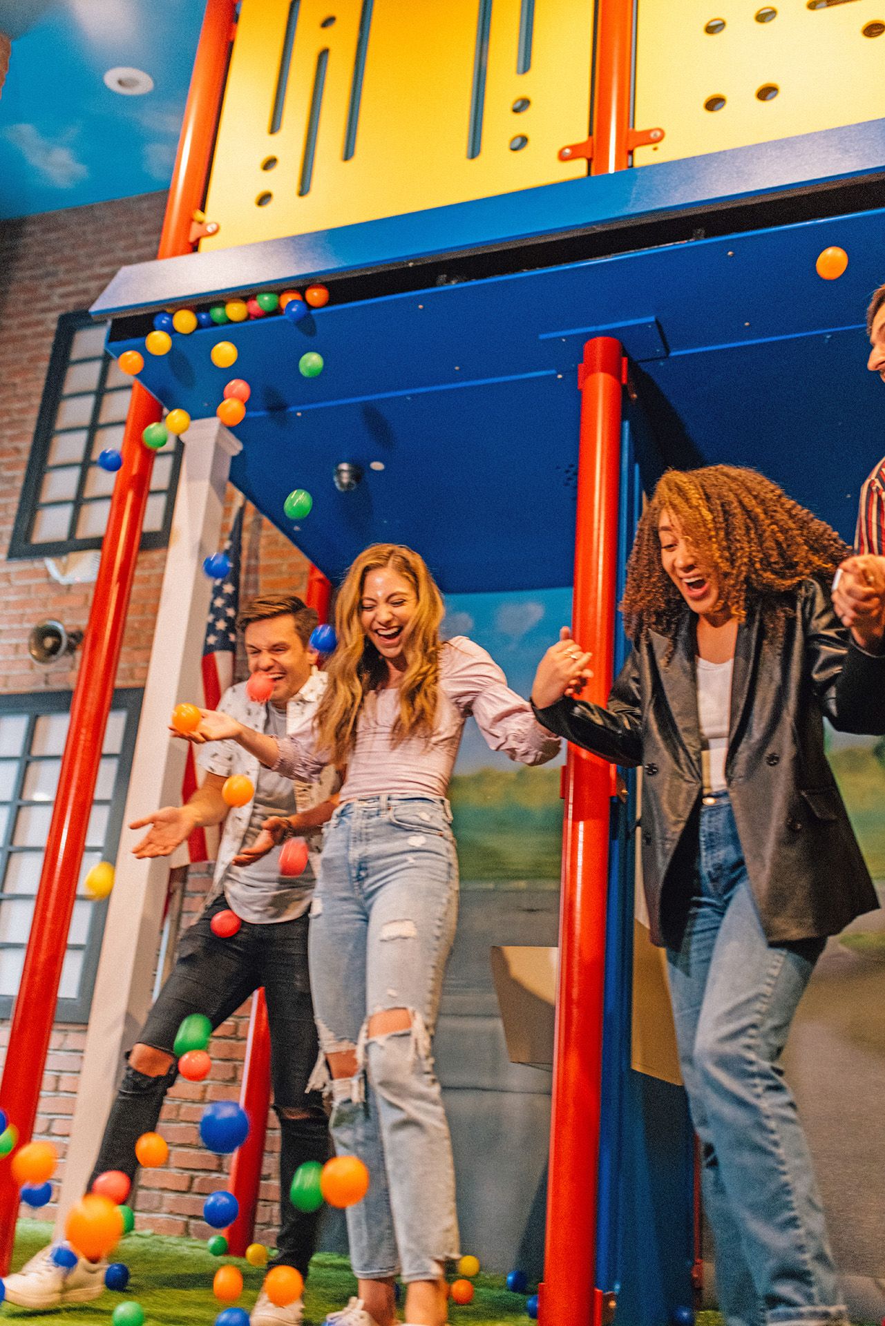 Three people laugh and play beneath a colorful play structure as balls cascade down around them.