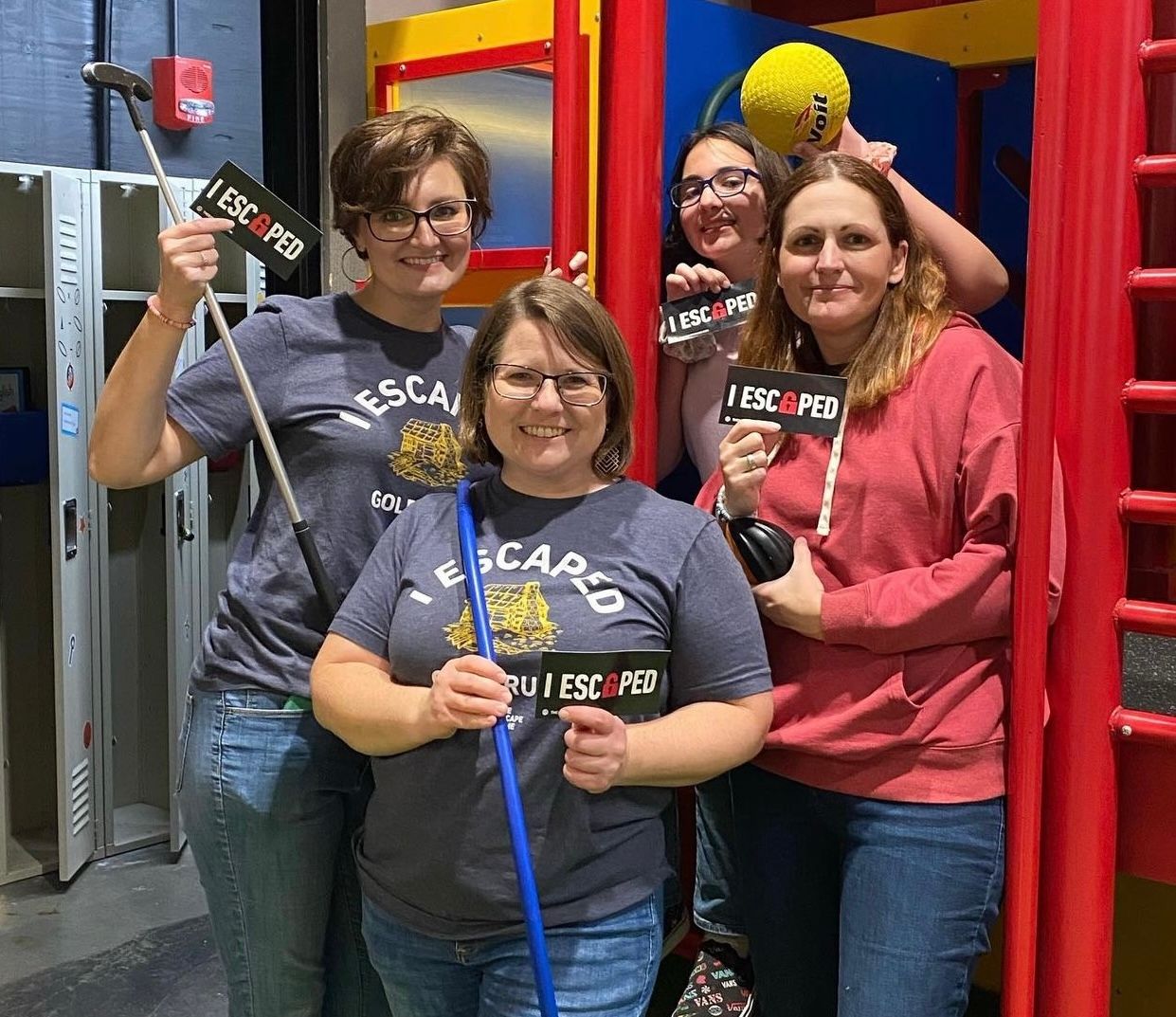 Four women stand together, smiling, holding signs that read "I ESCAPED." They pose in a colorful playground setting.