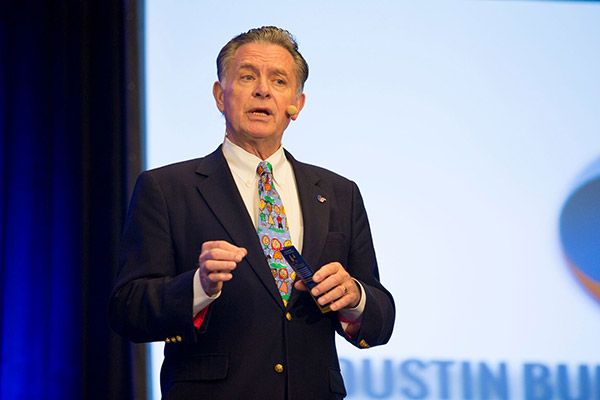 A man in a black suit with a colorful tie speaks at a podium, gesturing with his hands. A screen is visible behind him.