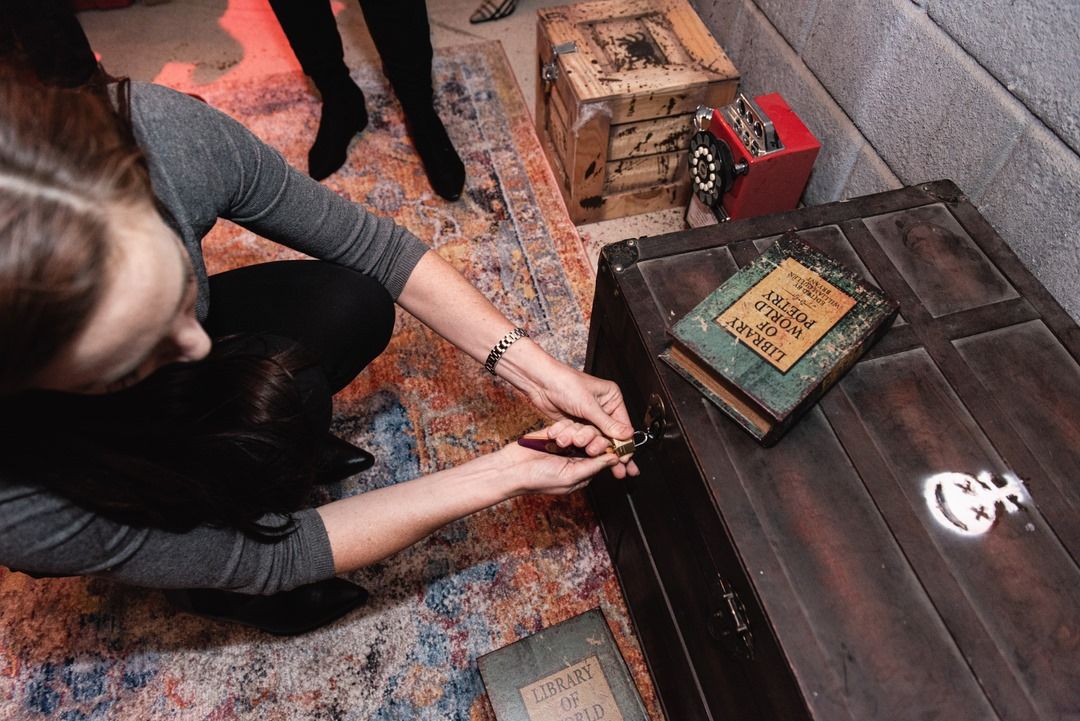 A person kneels, using a key to unlock a wooden chest with an old book titled "Library of Word Poetry" on top.