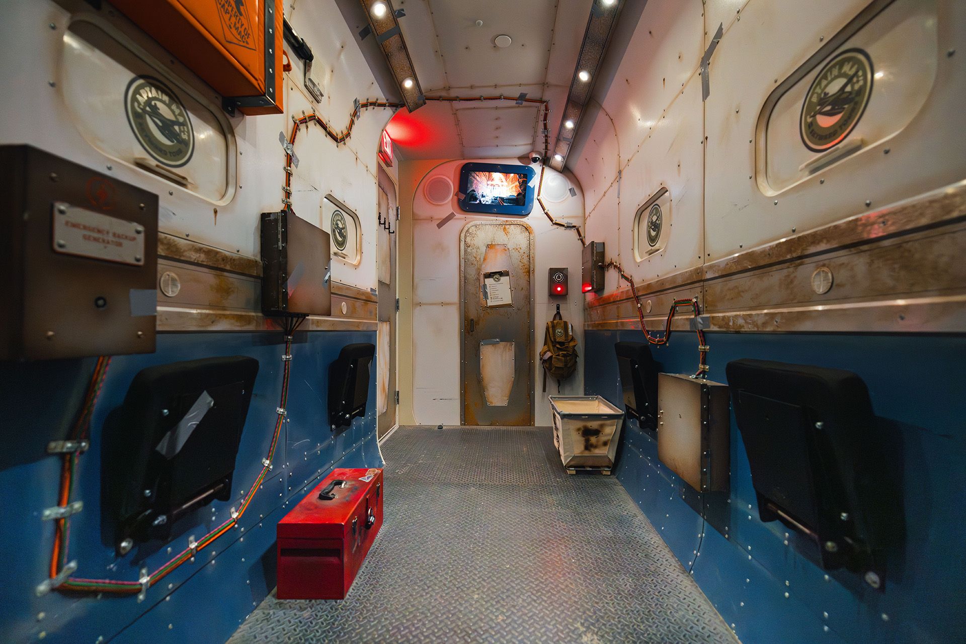 Interior of a futuristic, industrial corridor with metal walls, control panels, and seating along the sides.