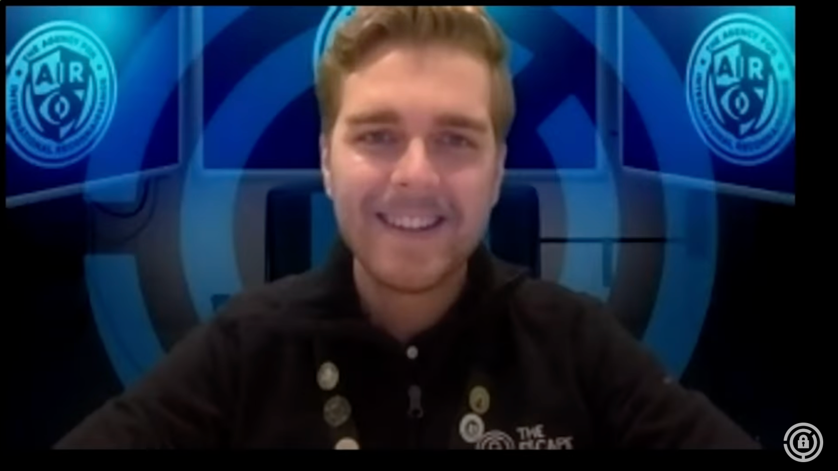 A young man with light brown hair smiles at the camera, wearing a black shirt with pins. A blue logo is in the background.