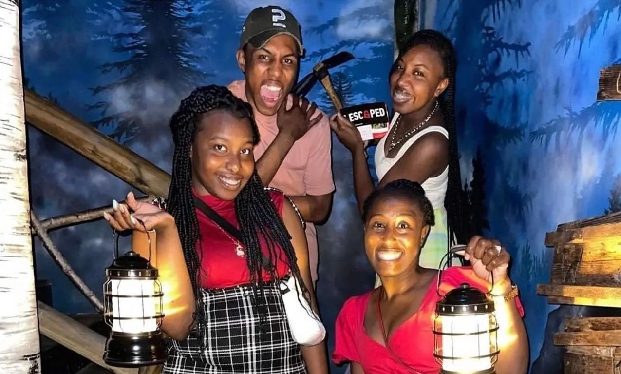 Four friends pose for a photo in a themed escape room, each holding lanterns. One person appears excited, holding a hammer.