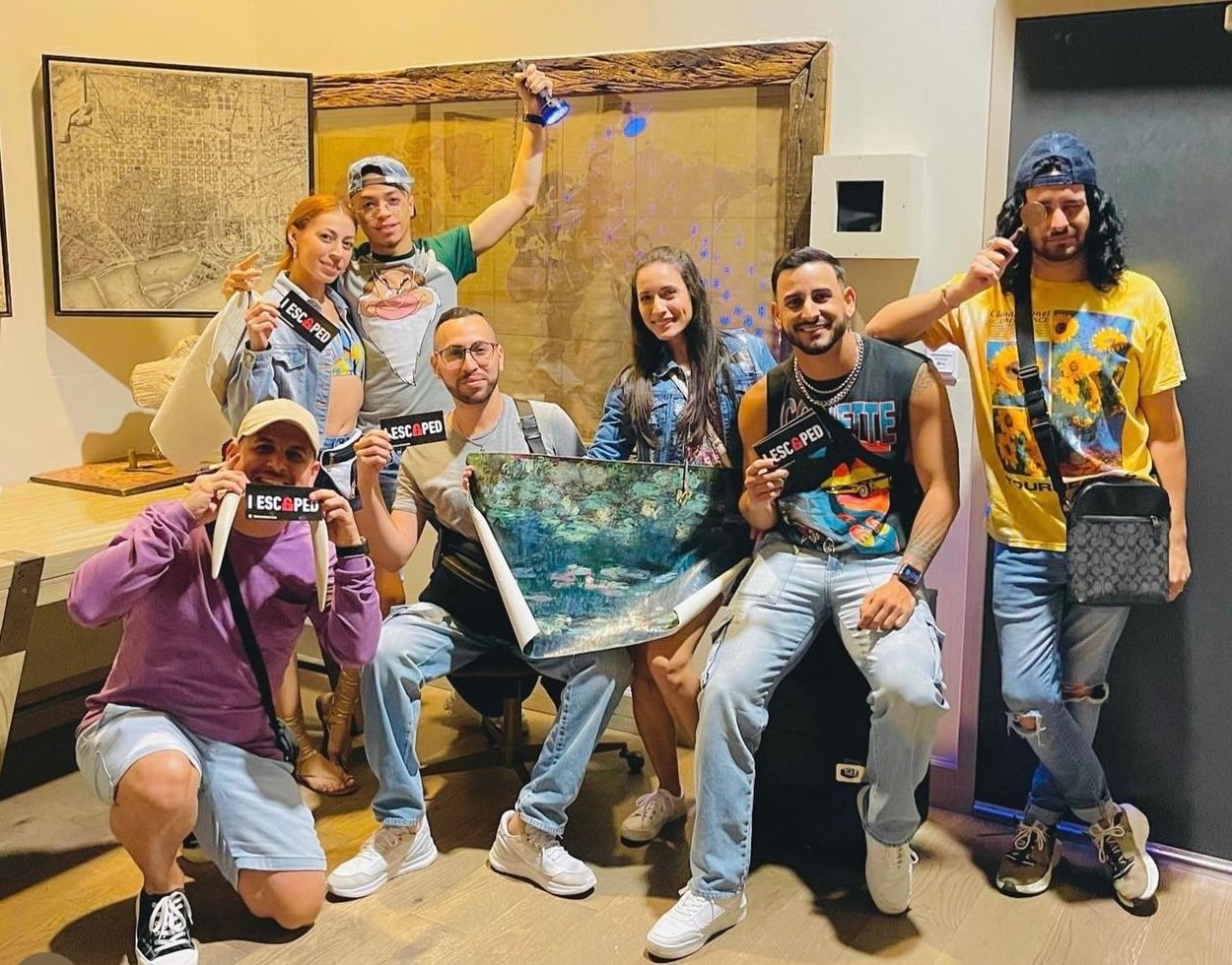 Group photo of seven people holding "ESCAPED" signs, smiling, with colorful clothing and a background featuring artwork.