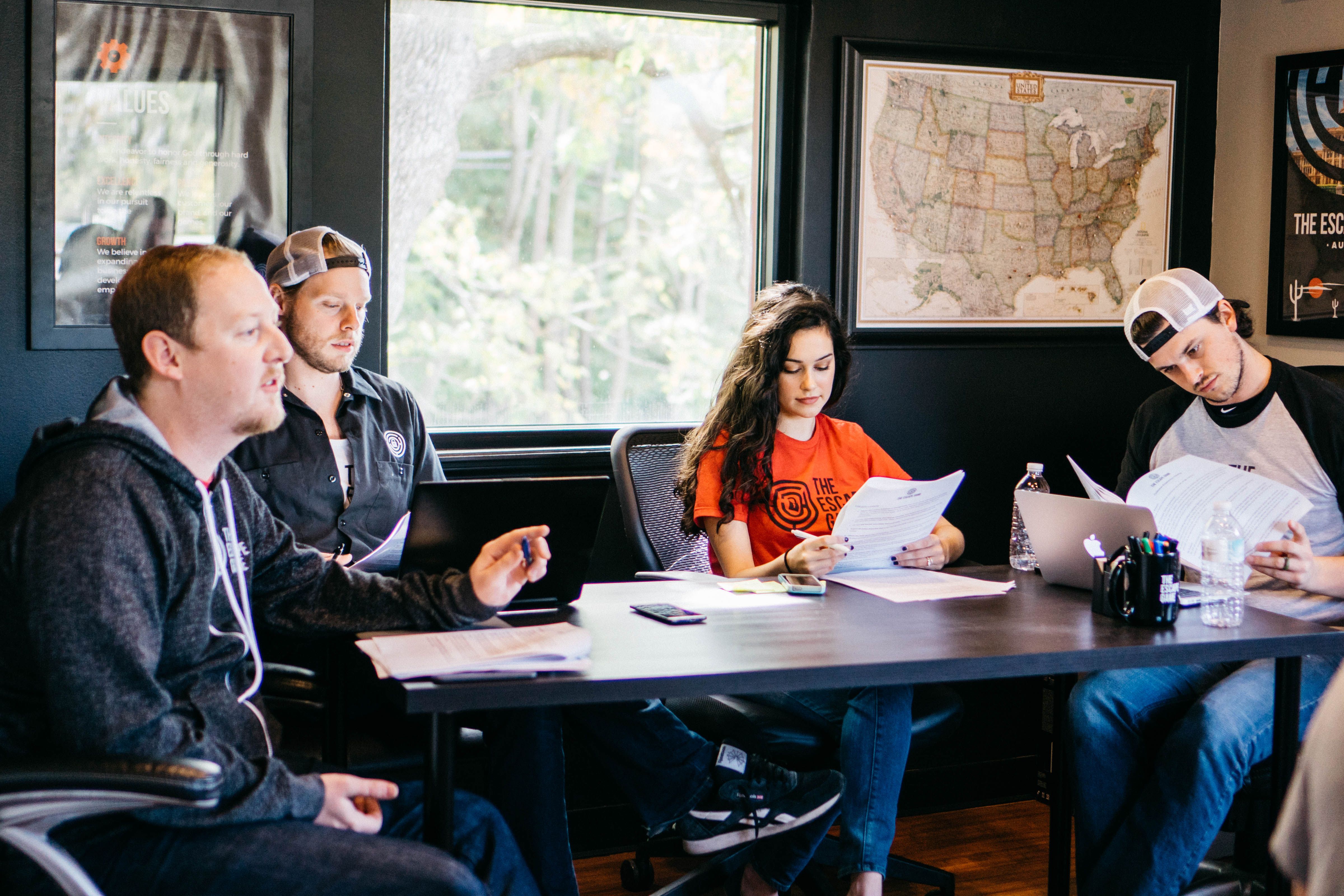Four individuals sit around a conference table, reviewing papers. A large map is on the wall, and there are windows with outside views.