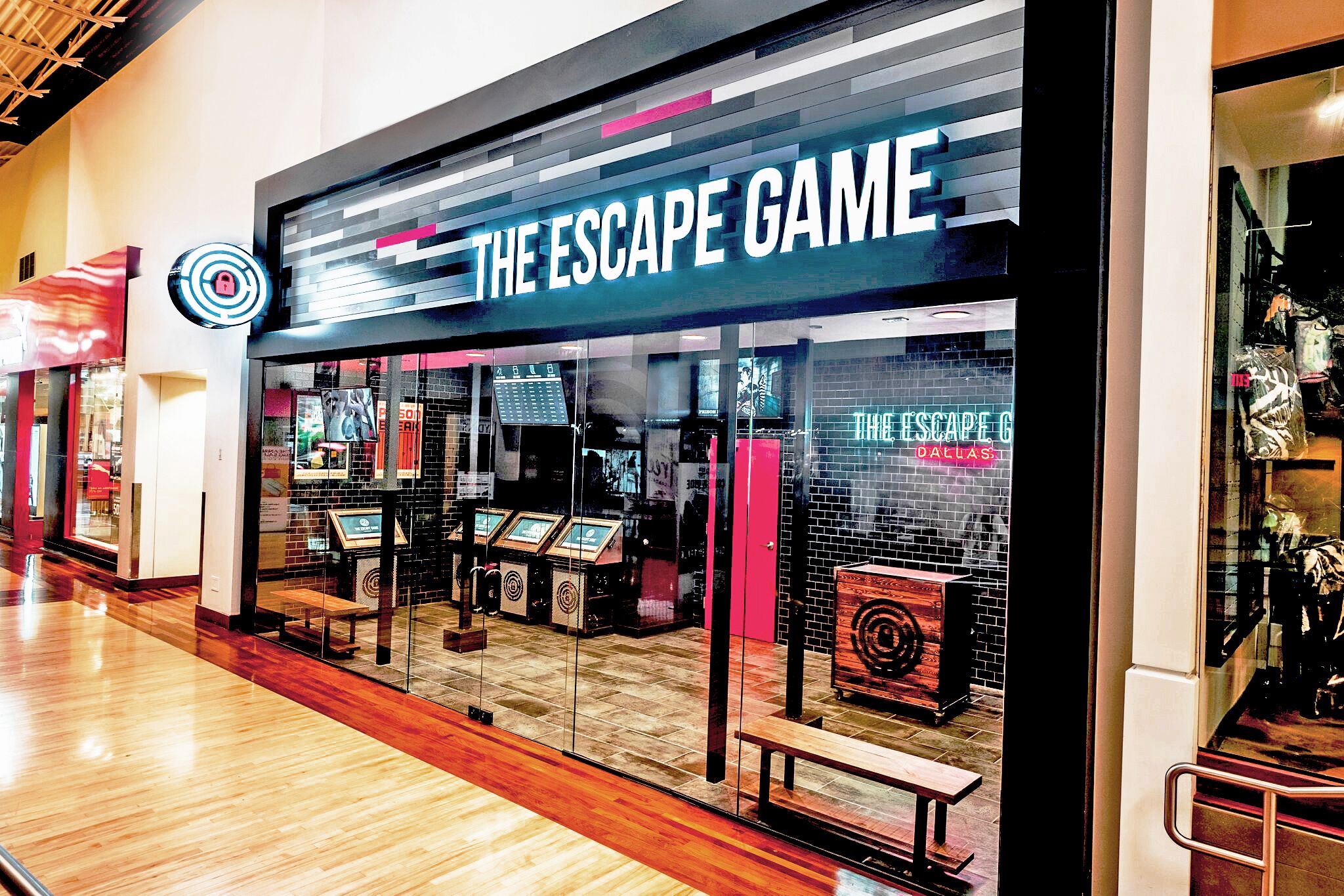 Exterior of "The Escape Game" with large signs, glass doors, displays, and a wooden bench in front. Bright and modern design.