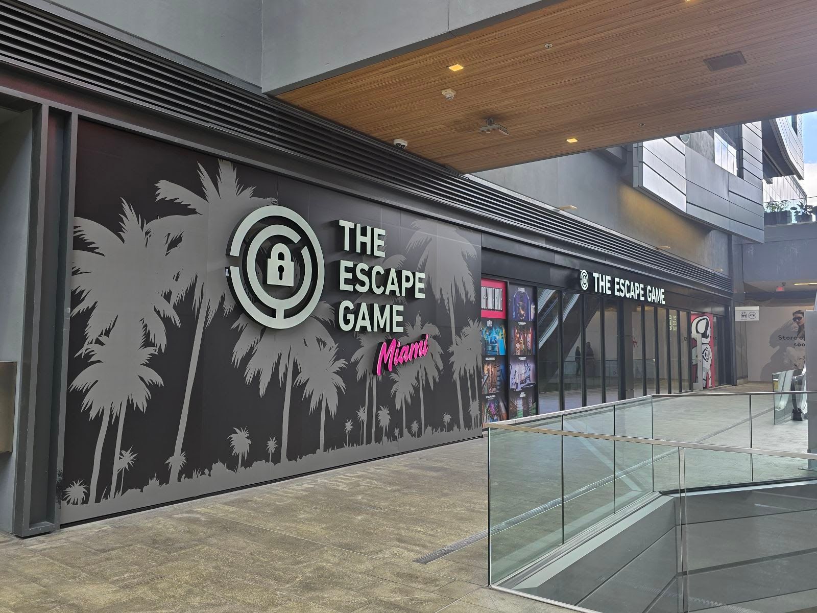 Entrance to "The Escape Game Miami," featuring palm trees and a logo, with glass railings and an open walkway.